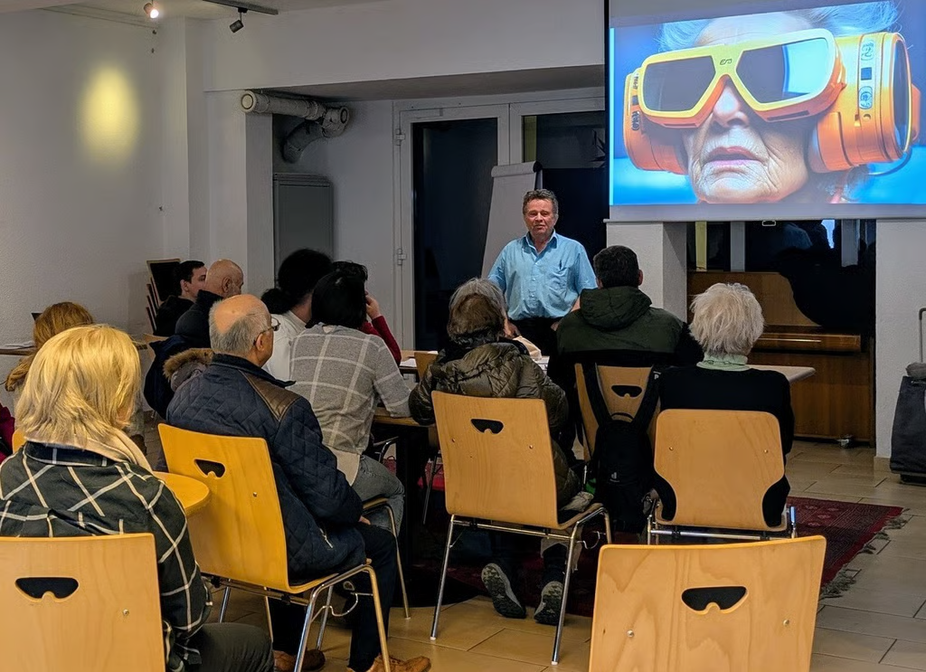 Ein Mann hält einen Vortrag vor einem kleinen Publikum in einem Seminarraum. Die Teilnehmenden sitzen auf Holzstühlen und schauen auf eine Leinwand, auf der das Bild einer älteren Person mit futurischer VR-Brille zu sehen ist. Die Szene vermittelt das Thema Digitalisierung oder virtuelle Realität im Bildungs- oder Informationskontext.