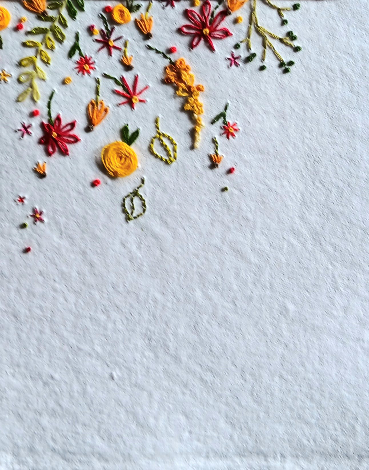 Detailaufnahme einer hellen Stofffläche mit feiner Stickerei: Oben sind bunte, florale Motive in Rot, Gelb, Orange und Grün gestickt – darunter verschiedene Blüten, kleine Zweige und Blätter. Der untere Bildbereich bleibt weitgehend leer und zeigt die schlichte, leicht strukturierte Stoffoberfläche.